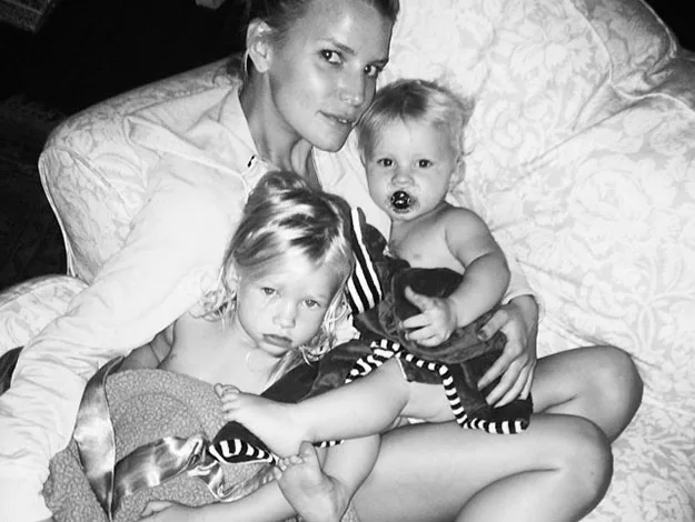 Woman seated on a couch, holding two young children, one with a pacifier, in a cozy, intimate setting.