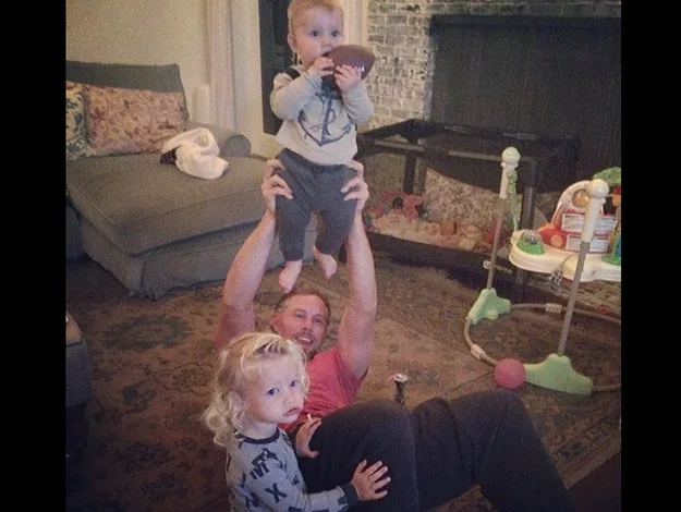 Man lying on the floor holding a baby up, with another child standing nearby in a cozy living room setting.