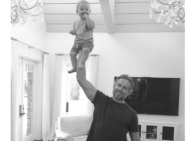 Man balancing a smiling toddler on his palm in a living room with chandeliers and a television in the background.