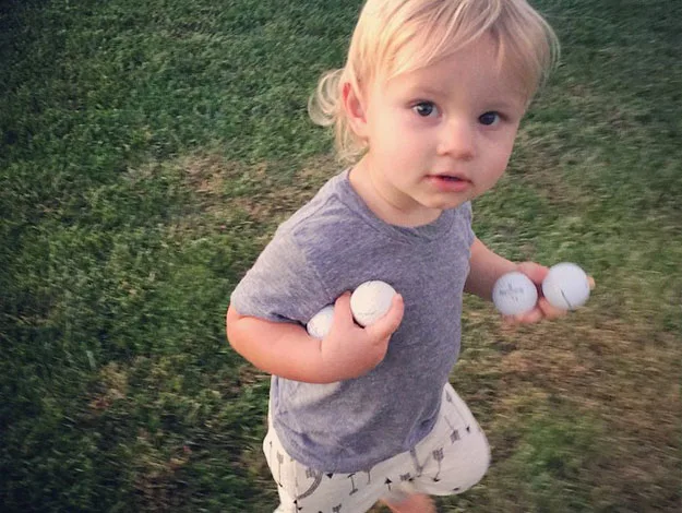 A toddler with blonde hair holds six golf balls while standing on grass.
