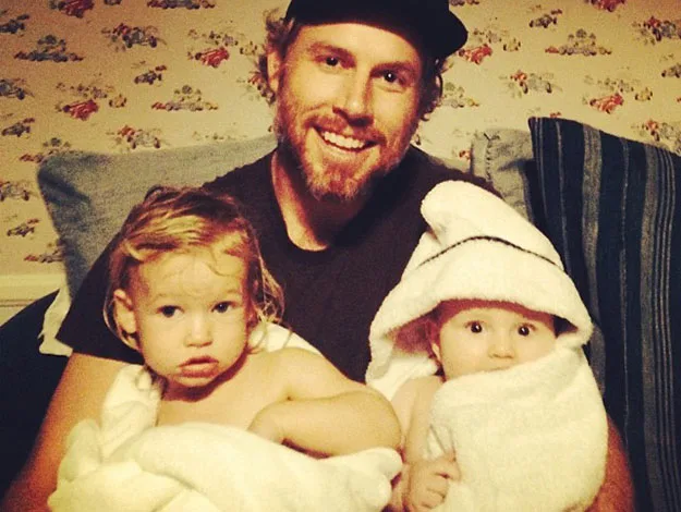 A bearded man smiling with two young children wrapped in towels on a couch.