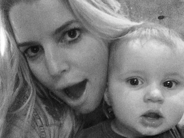 A woman with long, blonde hair poses playfully next to a baby, both with open mouths, in a black and white photo.