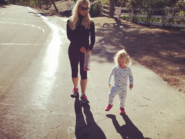 A woman and a toddler walking on a sunny path, with long shadows cast behind them.