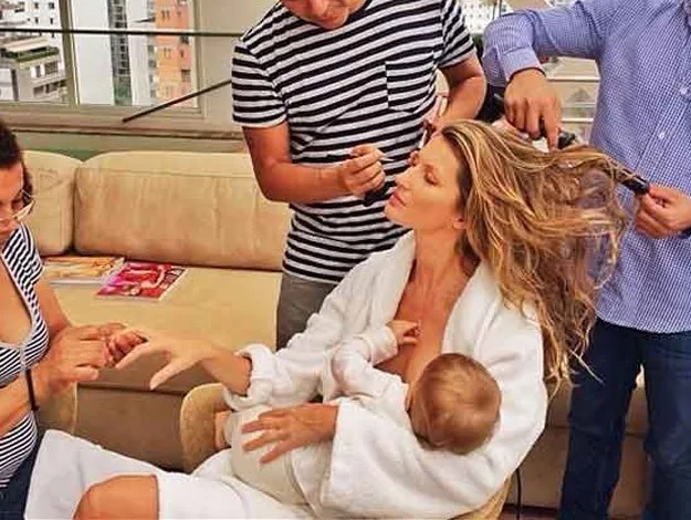 Woman in a white robe breastfeeding a baby while getting hair and nails done by stylists.