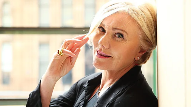 Woman with short blonde hair and a black jacket smiling while touching her hair, against a blurred urban background.
