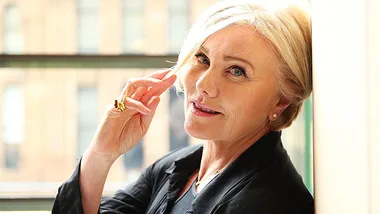 Woman with short blonde hair and a black jacket smiling while touching her hair, against a blurred urban background.