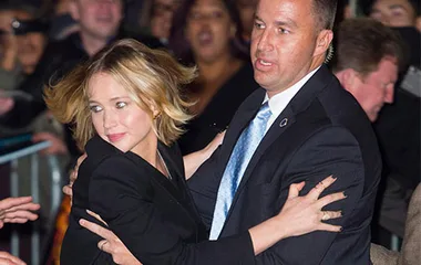A woman in a black outfit is being guided by a security guard through a crowd, looking concerned.