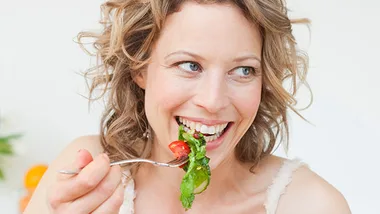 A woman happily eating a fresh salad with greens and cherry tomatoes on a fork.