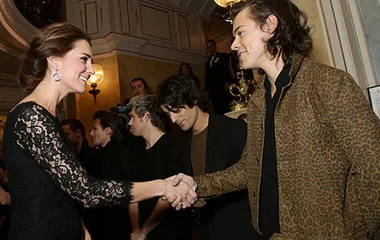 Royal greets band at event; woman in black lace shakes hands with man in leopard print, others in background observe.