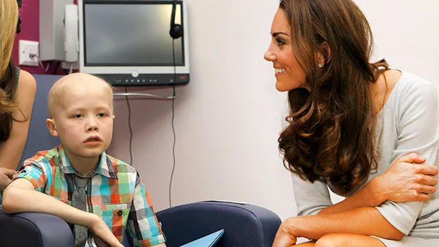 Duchess Catherine smiling at a young child with cancer, seated together in a hospital room setting.