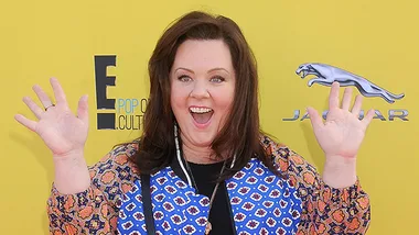 Melissa McCarthy smiling with hands raised, wearing a colorful outfit, standing against a yellow background with logos.
