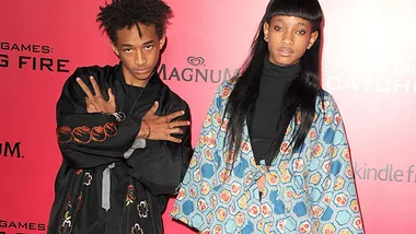 Jaden and Willow Smith pose on a red carpet wearing stylish outfits in front of a promotional backdrop.