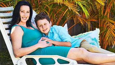 Woman in a green dress lounges on patio chair with a young boy, both smiling amidst lush greenery.