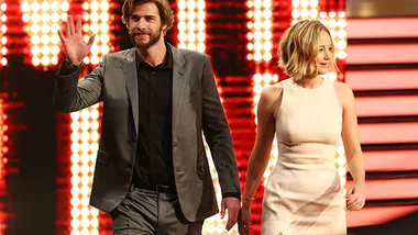 Two individuals walking on stage, one waving. Bright background lights and stylish attire hint at a formal event or show.