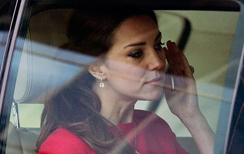 A woman in a red outfit seated in a car, appearing emotional, with one hand raised to her face.