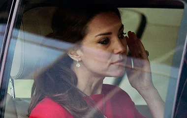 A woman in a red outfit seated in a car, appearing emotional, with one hand raised to her face.
