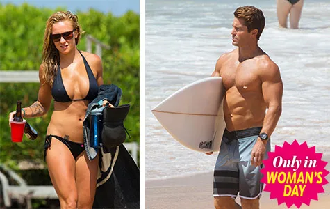 Man and woman on the beach; woman in a bikini holding a drink, man with a surfboard.