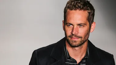 Man with a beard wearing a dark jacket, looking to the side against a plain background.
