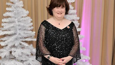 Woman in a sparkly black dress standing between white Christmas trees, smiling for a photo indoors.