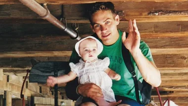 Father holding baby daughter, both smiling, under a rustic wooden ceiling.