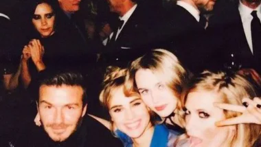 Four people smiling for a selfie at a social event; a woman in the background appears to be looking at the group.