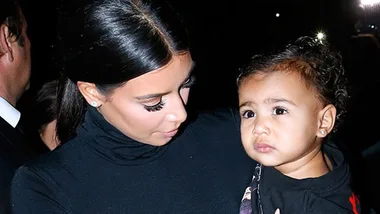 A woman, wearing a black outfit, holds a child at an event, illuminated by camera flashes in a dark setting.