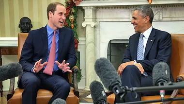 Prince William and President Obama smiling and chatting, seated in a formal setting with microphones in front.