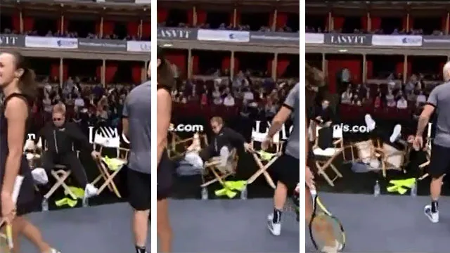 Elton John falls off chair at tennis match, players nearby react, indoor court with audience in background.
