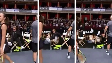 Elton John falls off chair at tennis match, players nearby react, indoor court with audience in background.