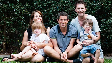 Family of five sitting together on grass, smiling, with greenery in the background.