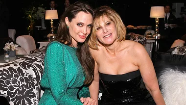 Two women sitting together at an event, one in a green dress and the other in a black dress, smiling at the camera.