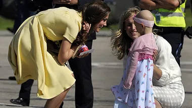 Duchess in yellow dress smiles and kneels to greet young girl with cancer, wearing a pink and white dress, held by woman.