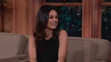 Mila Kunis smiling during a talk show interview, seated on a couch, wearing a black top.