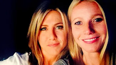 Two women smiling together, one with light hair and the other with medium hair, against a dark background.