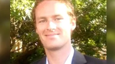 A smiling man in a suit stands outdoors with trees in the background.