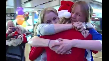 Three people embrace joyfully in an airport, one wearing a Santa hat, with balloons in the background.