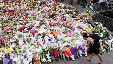 A woman places flowers among a large floral tribute on a city street, indicating a memorial or public mourning event.