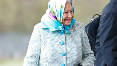 A smiling elderly woman with a blue floral scarf and coat stands outdoors, looking to her right.
