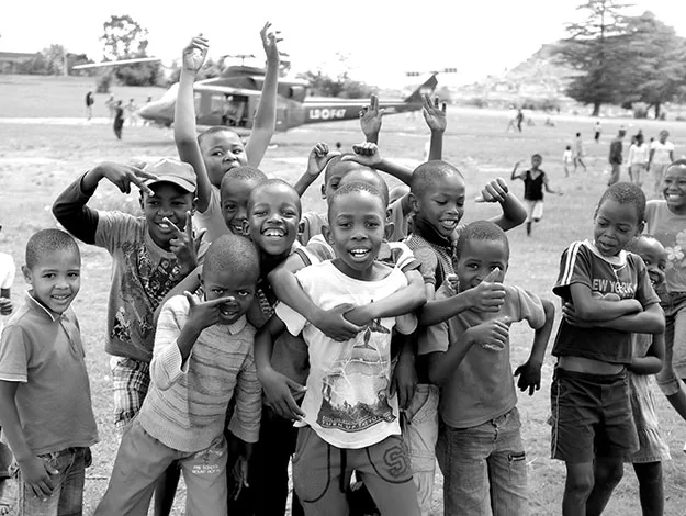 Group of smiling children posing outdoors in front of a helicopter, with some waving and others giving thumbs up.