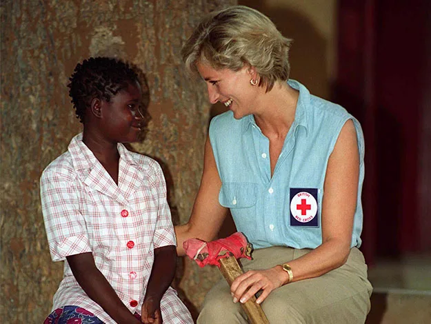 {"alt":"Woman with Red Cross badge smiling at a seated child against a tree."}