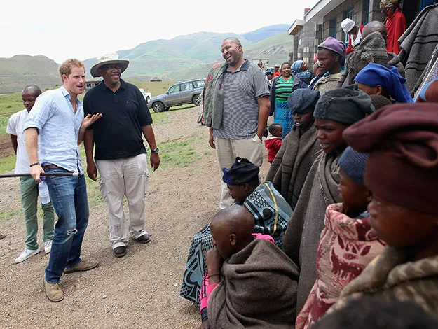 A group of people, including a man in a light blue shirt, smiling outdoors near a building, others wrapped in blankets.