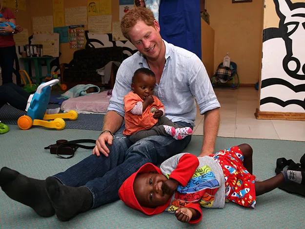 A man sitting on the floor, smiling, with a baby on his lap and another child lying nearby, in a playful setting.