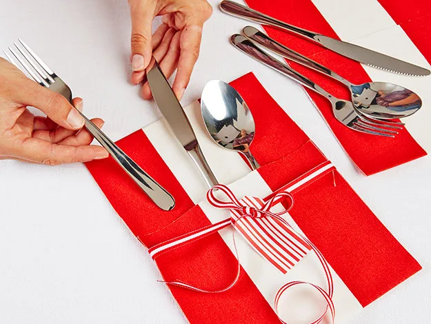 Red and white napkin wrapped cutlery tied with striped ribbon, hands arranging fork beside knife and spoon on white tablecloth.