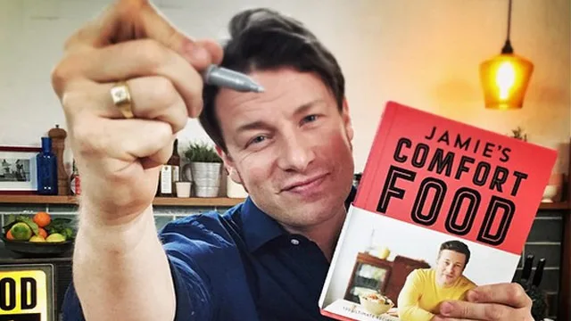 Chef holding a pen and his cookbook "Jamie's Comfort Food" in a kitchen setting.