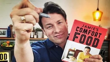 Chef holding a pen and his cookbook "Jamie's Comfort Food" in a kitchen setting.