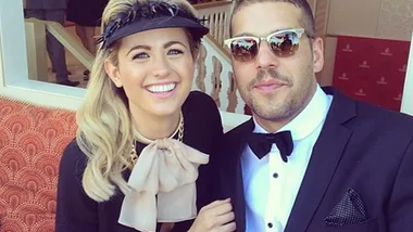 A couple dressed elegantly, smiling at a formal event; the woman wears a visor, the man a suit with sunglasses.