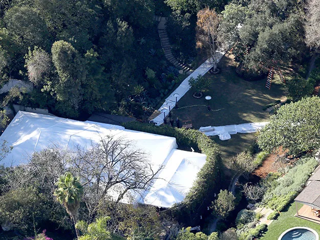 Aerial view of a large tent set up in a wooded backyard area, surrounded by trees and pathways.