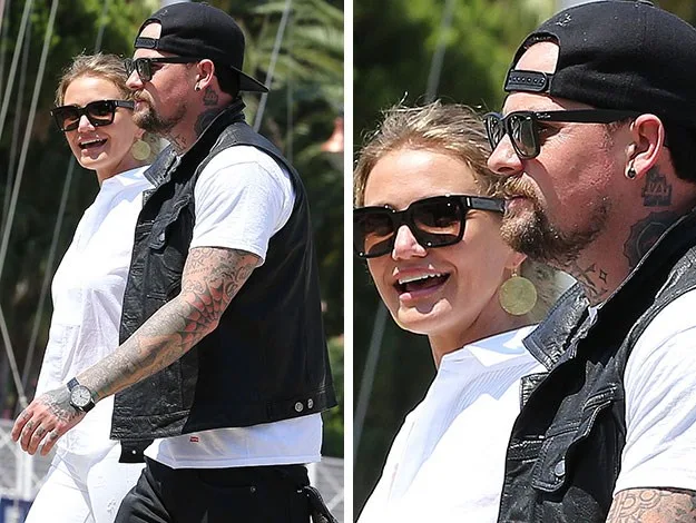 A man and woman wearing sunglasses walking together outdoors; the man has tattoos and a cap.