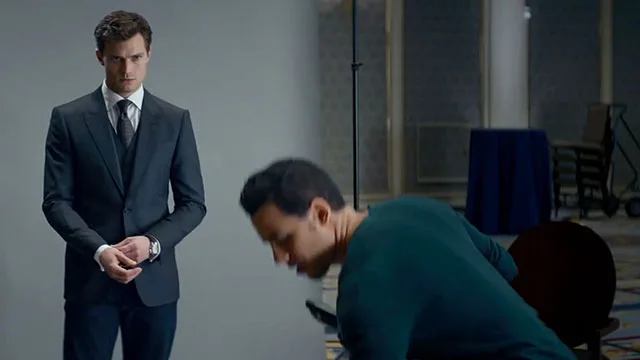 A suited man stands solemnly beside another bending man, set in an interior space with muted colors.