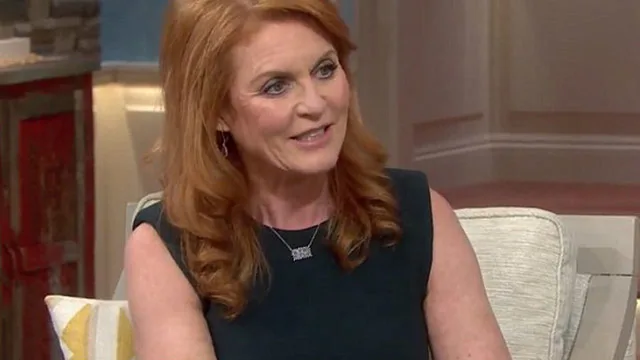 Sarah Ferguson smiling in a living room setting wearing a dark dress, seated on a light-colored sofa.
